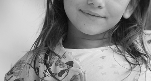 طفلة تتوسط صورة باللون الأسود والأبيض، ترتدي قميصاً مزين بزخارف نباتات، ويبدو نصف وجهها بابتسامة.
